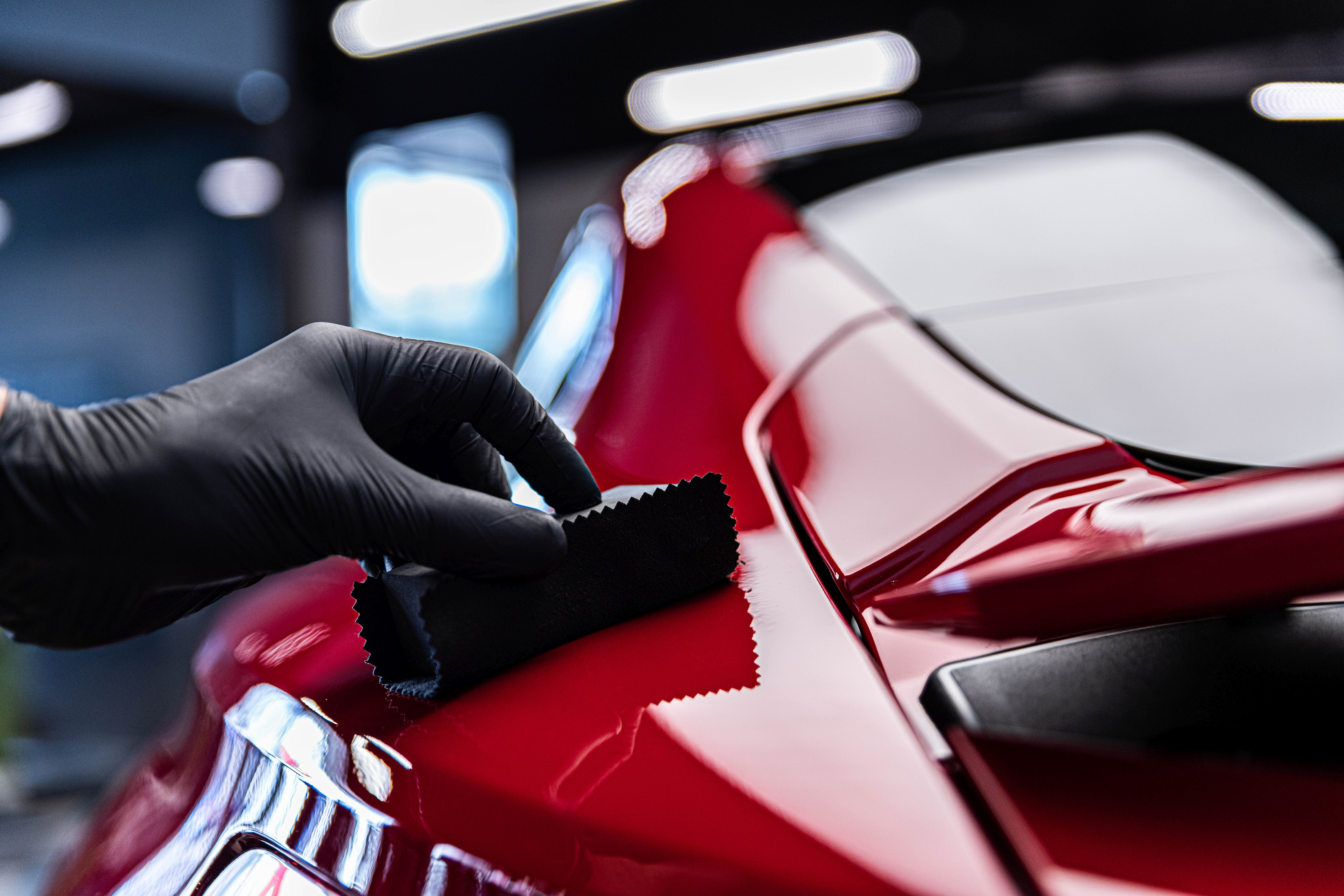Professional applying microfiber cloth to red car surface in showroom lighting